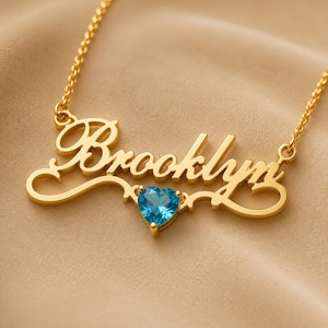 Op de afbeelding: Een goudkleurige naamketting met de naam Brooklyn in een cursief lettertype. Het ontwerp bevat een kleine, gefacetteerde, hartvormige blauwe edelsteen die onderaan in het midden van het naamplaatje is geplaatst, bevestigd aan een fijne gouden ketting.