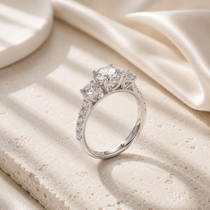 May include: A three-stone ring crafted from polished silver-toned metal, featuring a large round-cut center stone flanked by two smaller round-cut stones, with additional small round stones set along the upper portion of the ring band.