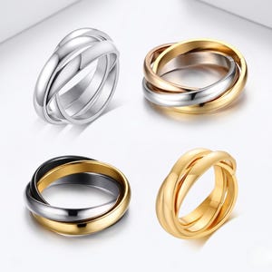 Könnte beinhalten: Vier Ringe in verschiedenen metallischen Ausführungen: Silber, Gold, Roségold und eine Kombination aus Gold und Schwarz. Jeder Ring hat ein Design mit drei Bändern, die miteinander verwoben sind. Die Ringe werden auf einer weißen Oberfläche präsentiert.