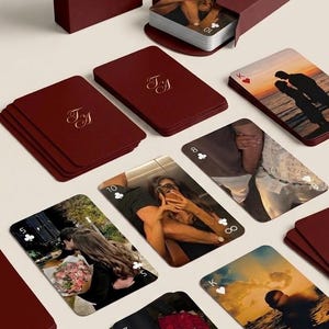 Op de afbeelding: Bordeaux speelkaarten met gouden monogram initialen in een bijpassende doos. De kaarten bevatten romantische foto's op de plaatjes. Een kaartspel wordt getoond met een doos en losse kaarten zijn verspreid.