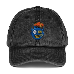 Puede incluir: Gorra de béisbol de mezclilla negra con un acabado desgastado. La gorra presenta una cara de monstruo azul bordada con detalles en amarillo y naranja. El monstruo tiene un ojo, un corazón y una boca sonriente.