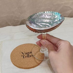 May include: Wine glass with an abalone shell bowl and a clear stem, held near a round cork coaster. The shell bowl has iridescent blues, greens, and silver hues. The coaster has the text "Vague Tether" printed on it.