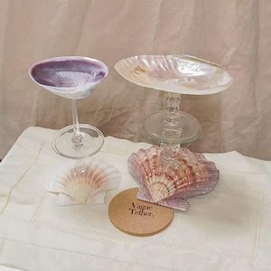 May include: Two seashell-based decorative items. One has a purple interior, the other is pearlescent. Both are mounted on clear glass stems and bases. A small, round coaster with the text "Vague Tether," is also present.