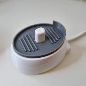 May include: A white and grey electric toothbrush charging base. The base is oval-shaped with a grey top featuring parallel grooves and a central white post. A white cable extends from the base.