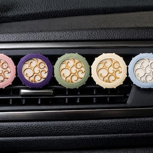 May include: Five colourful car air fresheners in pink, purple, green, cream, and blue. Each round freshener has a decorative gold-coloured swirl design in the centre. The fresheners are placed in the air vents of a car.