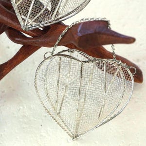 May include: Three heart-shaped ornaments crafted from fine silver-toned wire mesh. Each heart features a delicate metal chain attached to the top for hanging. The ornaments are suspended from a dark brown, branch-like wooden display stand.