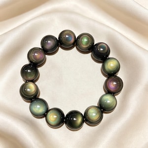 May include: A circular bracelet composed of smooth, polished spherical beads made of dark obsidian. Each bead features a distinct iridescent sheen, displaying concentric rings of green, gold, and purple hues that catch the light.