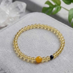 May include: A yellow beaded bracelet featuring small, translucent yellow beads. It includes a larger yellow bead and a small dark blue bead. The bracelet is presented on a grey surface.