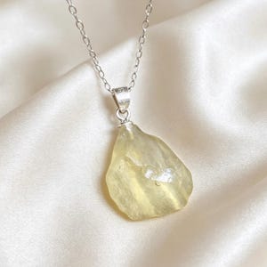 May include: A necklace featuring a raw, pale yellow gemstone pendant suspended from a delicate silver chain. The pendant has an irregular, organic shape with a translucent, polished surface, held by a simple silver bail stamped with the number 925.