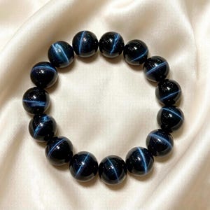 Op de afbeelding: Een armband bestaande uit glanzende, donkerblauwe, bolvormige kralen. Elke kraal heeft een kruisvormig, lichtblauw reflecterend patroon. De armband wordt gepresenteerd op een crèmekleurige, satijnachtige stof.