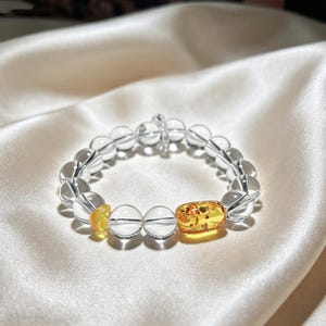 May include: A stretch bracelet composed of clear, spherical crystal beads. The design features one small, irregular yellow amber bead and one larger, rectangular, translucent yellow amber bead with visible internal inclusions.