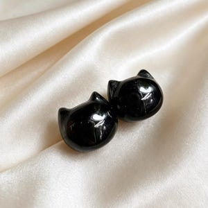 Könnte beinhalten: Ein Paar kleiner, glatter Ohrstecker in Form von schwarzen Katzenköpfen mit spitzen Ohren. Die Ohrringe haben eine glänzende, reflektierende Oberfläche und ein abgerundetes, kompaktes Design, das das Licht auf dem dunklen Finish einfängt.