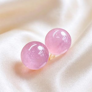 Peut inclure: Une paire de boucles d'oreilles à clous composée de deux pierres précieuses sphériques, lisses et polies, d'un rose clair. Chaque pierre ronde présente une surface brillante et réfléchissante avec de subtiles variations internes.