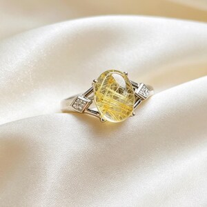 May include: A silver-toned ring featuring a large, oval-shaped rutilated quartz gemstone with visible golden needle-like inclusions. The band is accented on each side by a small, square-shaped setting containing four tiny, clear, sparkling stones.