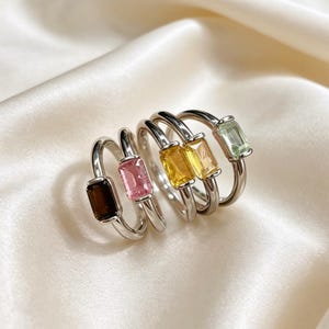 May include: A collection of five silver-toned rings, each featuring a single rectangular baguette-cut gemstone. The stones are arranged in a row and display a variety of colours including dark brown, light pink, two shades of yellow, and pale green.