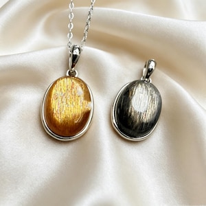 Può includere: Due ciondoli ovali con montature in metallo color argento. Un ciondolo presenta una pietra scintillante color oro-arancio, mentre l'altro contiene una pietra grigio scuro con venature metalliche. Il ciondolo dorato è fissato a una sottile catenina argentata.