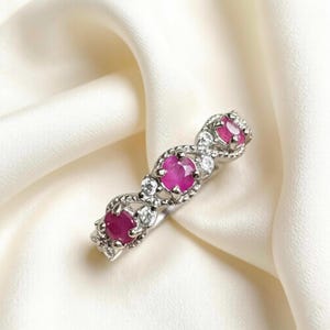 Può includere: Un anello d'argento con tre pietre preziose rosse ovali, impreziosito da pietre chiare più piccole. L'anello è posto su uno sfondo bianco morbido, che mette in risalto il design e il colore del gioiello.