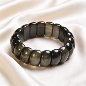 May include: A stretch bracelet composed of smooth, rectangular, polished beads made of dark, translucent stone with golden, shimmering inclusions throughout the surface. The beads are arranged in a continuous circular loop, reflecting light.
