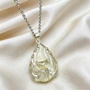 May include: A pale yellow, translucent, teardrop-shaped pendant featuring intricate, carved, swirling patterns. The pendant is suspended from a silver-toned metal chain with oval links, creating a cohesive piece of jewellery with a polished finish.