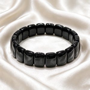 Op de afbeelding: Een elastische armband bestaande uit gladde, rechthoekige, gepolijste zwarte kralen. De kralen zijn gerangschikt in een doorlopende cirkelvormige lus en hebben een hoogglanzende afwerking die het licht op hun donkere oppervlakken weerkaatst.