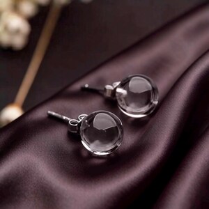 May include: A pair of clear, spherical stud earrings with silver posts. The earrings are presented on a dark purple satin fabric, which contrasts with their transparency. These earrings are a simple, elegant accessory.
