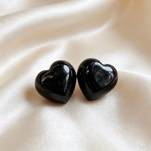 Könnte beinhalten: Ein Paar Ohrstecker mit glatten, glänzenden, schwarzen herzförmigen Elementen. Die Herzen haben ein abgerundetes, dreidimensionales, gewölbtes Design mit einer reflektierenden Oberfläche, die das Licht einfängt und subtile Glanzlichter auf der dunklen, polierten Oberfläche erzeugt.