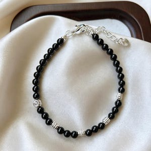 Könnte beinhalten: Schwarzes Perlenarmband mit silberfarbenen Akzenten. Das Armband besteht aus kleinen, runden schwarzen Perlen und dekorativen silberfarbenen Perlen, mit einem silbernen Verschluss. Das Armband wird auf einer weißen Satin-Oberfläche präsentiert.