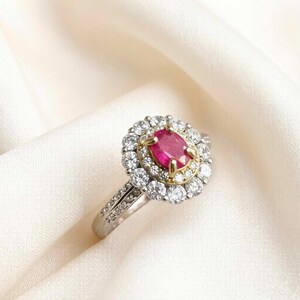 May include: A silver ring featuring a central oval ruby, encircled by a halo of small, clear gemstones. The ruby is a vivid red, with a gold accent around it. The ring's band is also embellished with smaller gemstones, creating a sparkling effect.