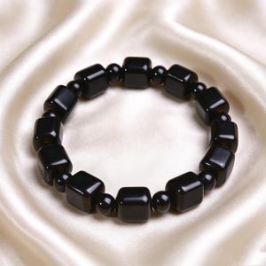 Könnte beinhalten: Ein Armband bestehend aus abwechselnd schwarzen würfelförmigen Perlen und kleineren schwarzen kugelförmigen Perlen, die in einem Kreis angeordnet sind. Die Perlen haben eine glatte, polierte Oberfläche und sind zu einer flexiblen Schlaufe aufgereiht.