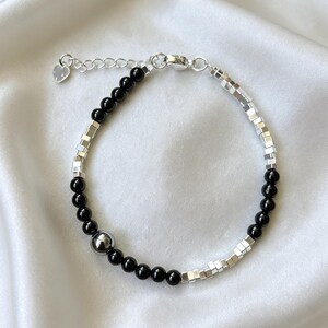 Könnte beinhalten: Ein silbernes Armband mit abwechselnden Abschnitten aus kleinen, quadratischen Silberperlen und polierten schwarzen Perlen. Eine größere, silberfarbene Perle ist mittig angeordnet. Die verstellbare Kette hat einen herzförmigen Anhänger.