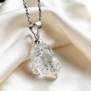 Op de afbeelding: Een heldere kwartskristallen hanger met een zilveren ketting. Het kristal heeft een onregelmatige vorm met interne insluitsels en is opgehangen aan een zilveren balein. De achtergrond is een witte, zijdeachtige stof.
