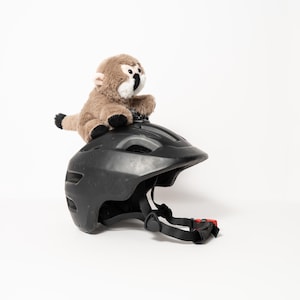 May include: A black bicycle helmet with a red buckle and straps. A small stuffed monkey with brown fur and black accents sits on top of the helmet. The helmet is designed for safety and protection.
