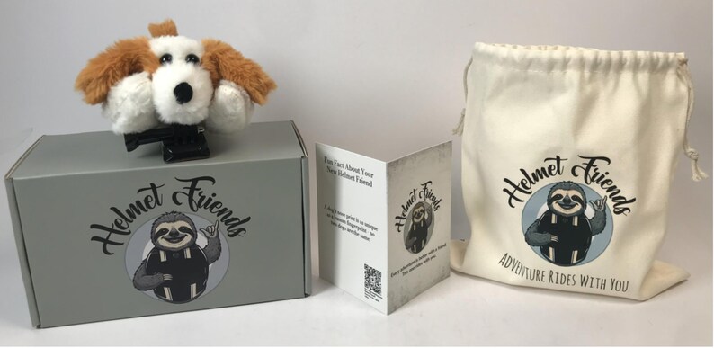 May include: A plush dog helmet cover with brown and white fur sits atop a gray box. The box and a drawstring bag feature the "Helmet Friends" logo with a sloth. A folded card is also included.