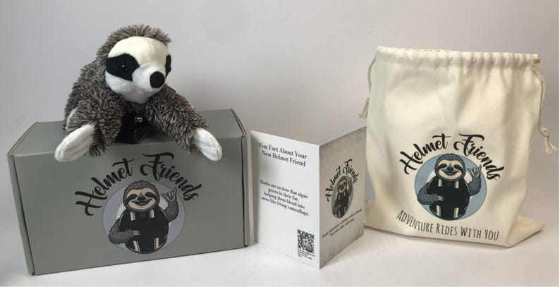 May include: A plush sloth toy sits atop a gray box with the words "Helmet Friends" and a sloth illustration. A drawstring bag and a folded card with the same logo are also present. The bag reads "Adventure Rides With You."