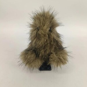 May include: A brown and black faux fur pom-pom with a black base. The pom-pom has long, spiky hairs and a rounded shape. The base is rectangular. The item is set against a white background.