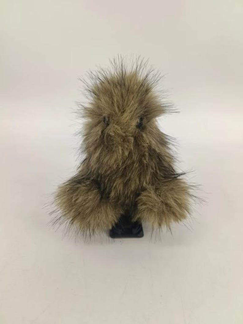 May include: A brown and black faux fur pom-pom with a black base. The pom-pom has long, spiky hairs and a rounded shape. The base is rectangular. The item is set against a white background.