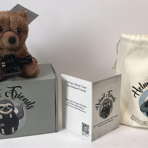 May include: A brown plush bear toy with a black accessory sits atop a gray box. The box and a drawstring bag feature the "Helmet Friends" logo with a sloth illustration. A folded card provides information.