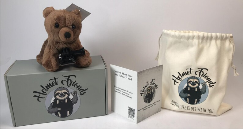 May include: A brown plush bear toy with a black accessory sits atop a gray box. The box and a drawstring bag feature the "Helmet Friends" logo with a sloth illustration. A folded card provides information.