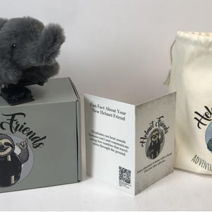 May include: A gray plush elephant toy sits atop a gray box with the words "Helmet Friends" and a sloth logo. A drawstring bag and a folded card with the same logo are also present. The bag reads "Adventure Rides With You."