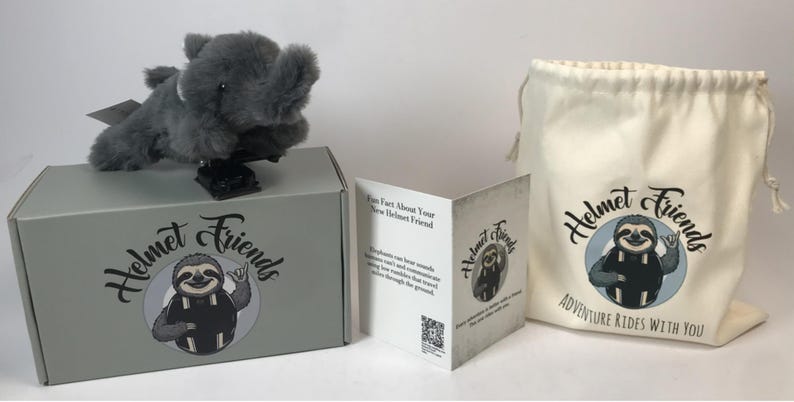 May include: A gray plush elephant toy sits atop a gray box with the words "Helmet Friends" and a sloth logo. A drawstring bag and a folded card with the same logo are also present. The bag reads "Adventure Rides With You."