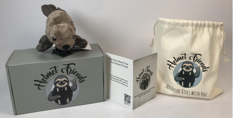 May include: A plush seal toy sits atop a gray box with the "Helmet Friends" logo. A drawstring bag and a folded card with the same logo are also present. The bag reads "Adventure Rides With You."