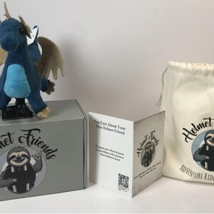 May include: A plush blue dragon with gold glitter wings and accents sits atop a gray box. The box and a drawstring bag feature the "Helmet Friends" logo with a sloth. A folded card is also included.