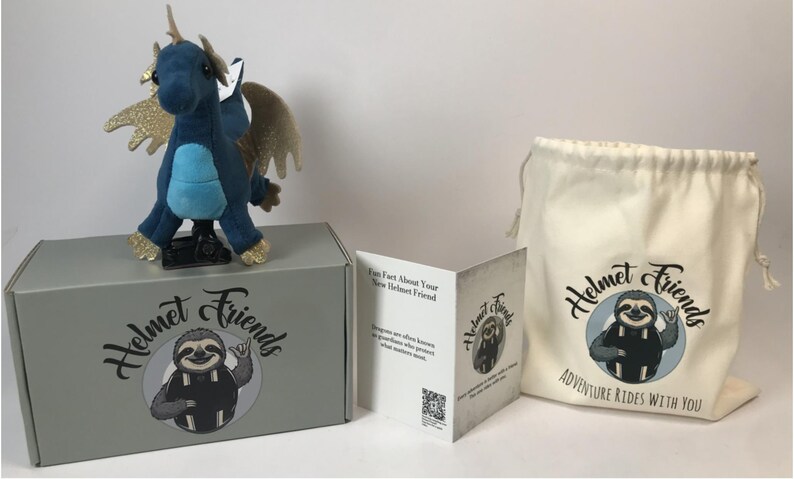May include: A plush blue dragon with gold glitter wings and accents sits atop a gray box. The box and a drawstring bag feature the "Helmet Friends" logo with a sloth. A folded card is also included.