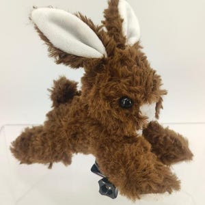 May include: A brown plush bunny with white inner ears and a white patch on its face. The bunny has black eyes and a fluffy tail. It is lying down and appears to be attached to a black mount.