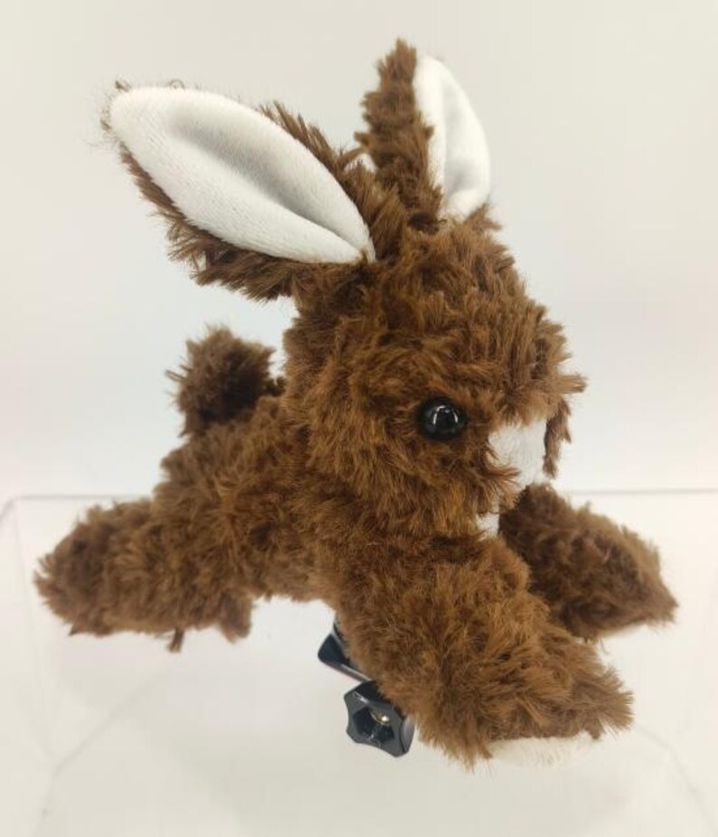 May include: A brown plush bunny with white inner ears and a white patch on its face. The bunny has black eyes and a fluffy tail. It is lying down and appears to be attached to a black mount.