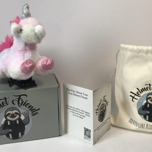May include: A plush unicorn toy with a silver horn and pink mane and hooves sits atop a gray box. The box and a drawstring bag feature a sloth logo with the text "Helmet Friends". A folded card is also present.