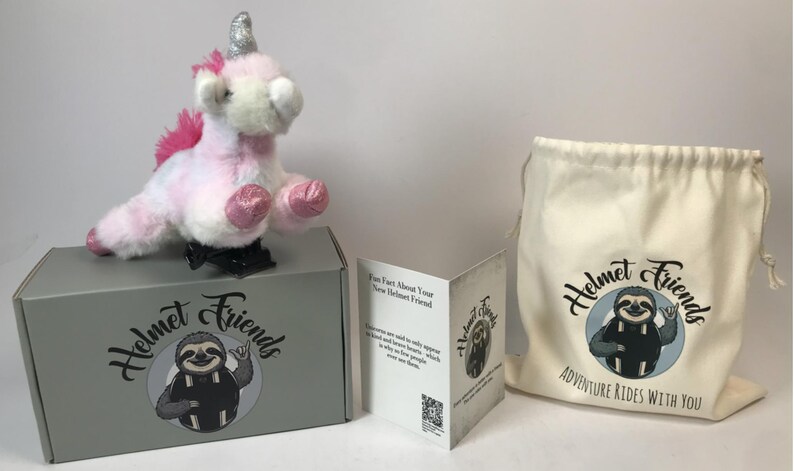 May include: A plush unicorn toy with a silver horn and pink mane and hooves sits atop a gray box. The box and a drawstring bag feature a sloth logo with the text "Helmet Friends". A folded card is also present.