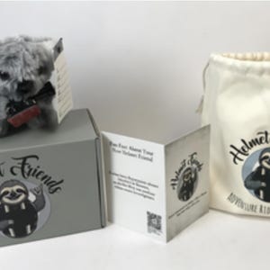 May include: A plush koala toy with a black bow tie sits atop a gray box, next to a drawstring bag and a folded card. The box, bag, and card feature the "Helmet Friends" logo with a sloth illustration. The bag reads "Adventure Rides With You."