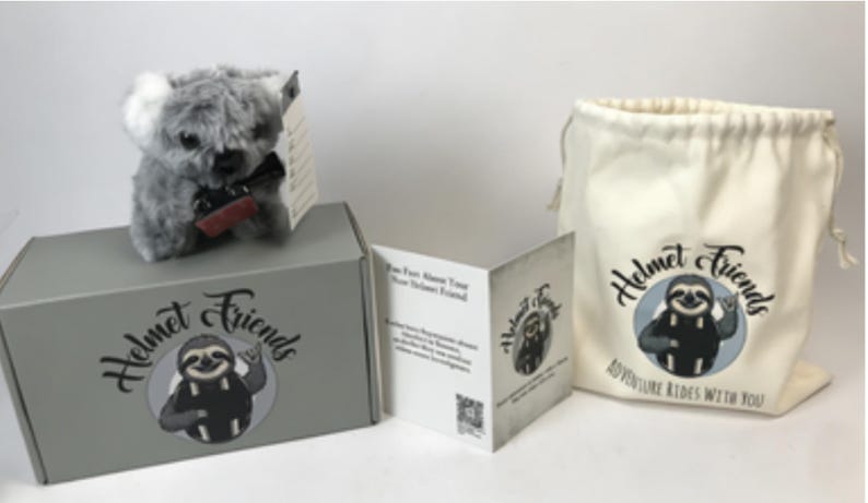 May include: A plush koala toy with a black bow tie sits atop a gray box, next to a drawstring bag and a folded card. The box, bag, and card feature the "Helmet Friends" logo with a sloth illustration. The bag reads "Adventure Rides With You."
