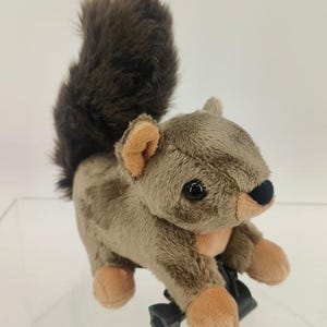 May include: A plush squirrel toy with a fluffy brown tail and tan body. The toy has a black nose, black eyes, and tan paws and ears. The squirrel is attached to a black plastic clip.
